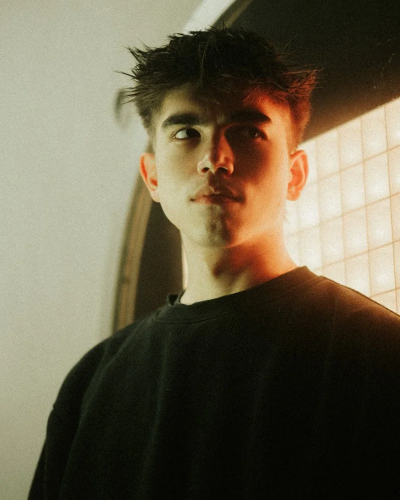 Junger Mann mit dunklen, zerzausten Haaren, trägt schwarzen Pullover, halb im Schatten, im Profil gesehen.