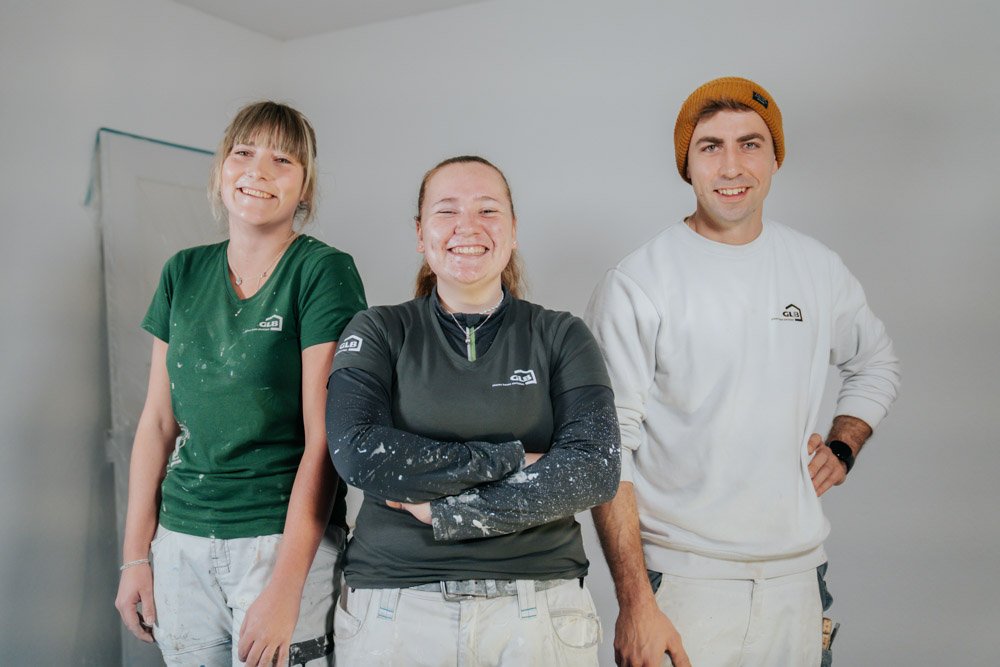 Drei lachende Menschen in Arbeitshosen und T-Shirts mit einem Logo stehen vor einer weißen Wand, auf einer Baustelle.