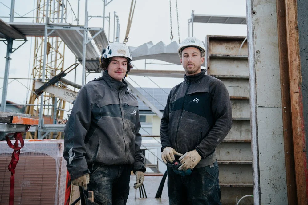 Zwei Bauarbeiter mit Helmen stehen auf einer Baustelle, umgeben von Gerüsten und Baumaterialien.