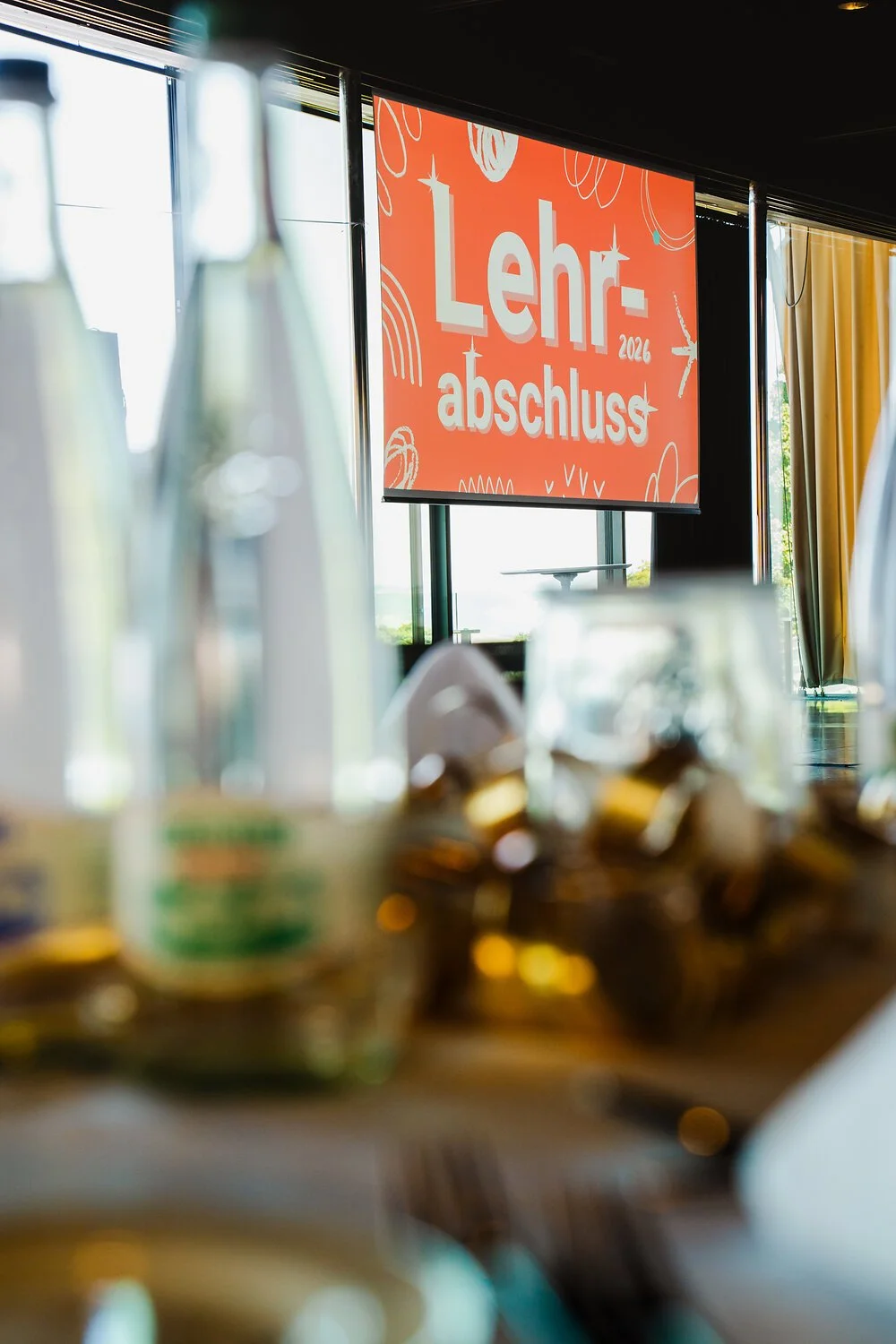 Bild von einem Saal mit Tischen, auf denen Flaschen und Gläser stehen, im Hintergrund ein Bildschirm mit der Aufschrift "Lehrabschluss 2026" in rot mit weissen Buchstaben.