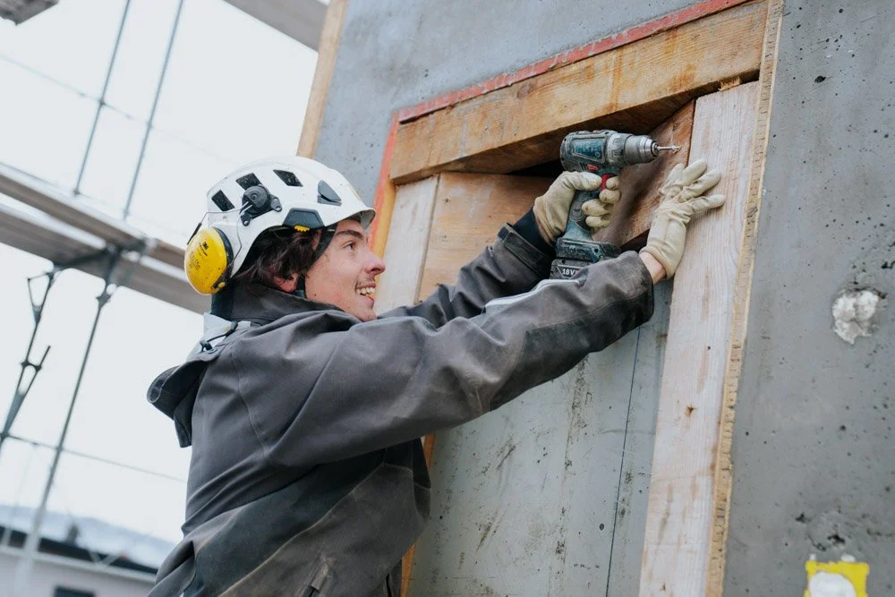 Ein Bauarbeiter trägt einen Schutzhelm und arbeitet mit einem Akku-Bohrer an einer Holzöffnung in eine Betonwand.
