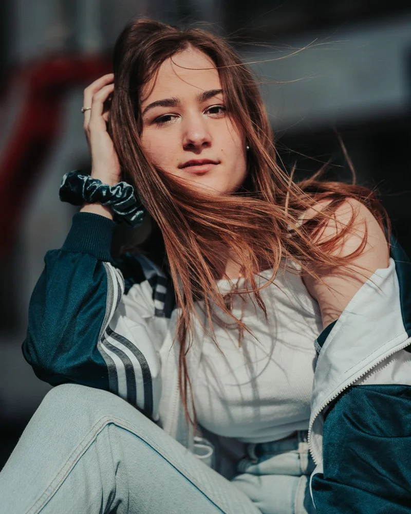 Junge Frau mit langen braunen Haaren, die über der Schulter hängen, trägt eine Jacke und ein T-Shirt.