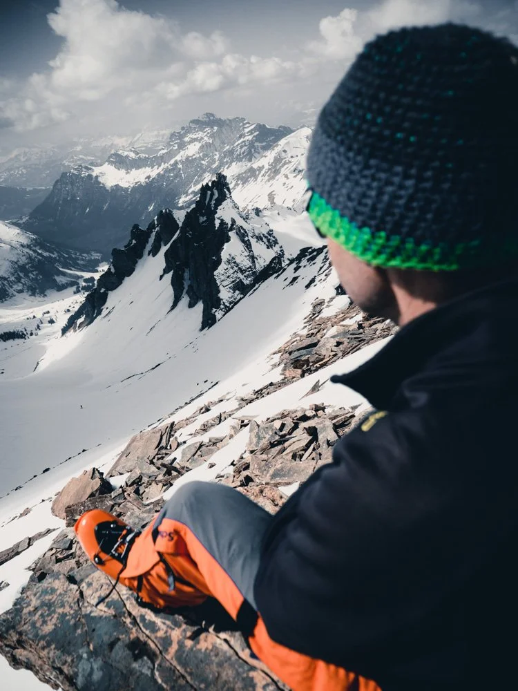 Ein Bergsteiger sitzt auf einem Gestein in schneebedeckten Bergen, trägt eine dunkle Jacke, eine bunte Mütze und hält eine orange Schneeschaufel.
