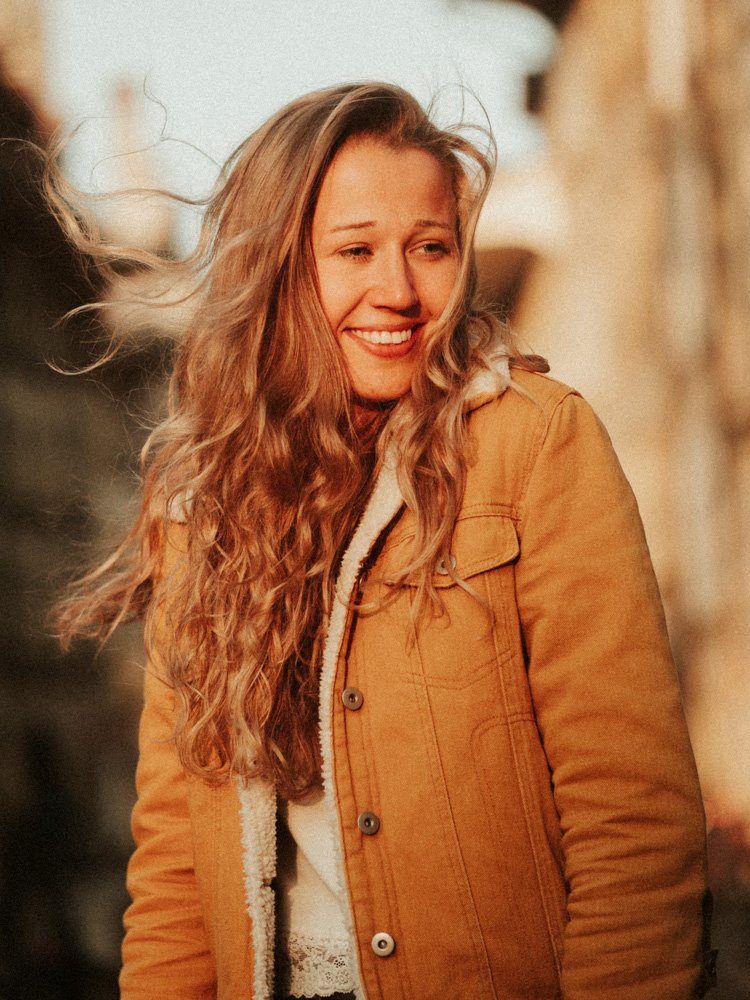 Junge Frau mit langen, lockigen Haaren lächelnd, trägt braune Jacke, im Freien bei Sonnenuntergang