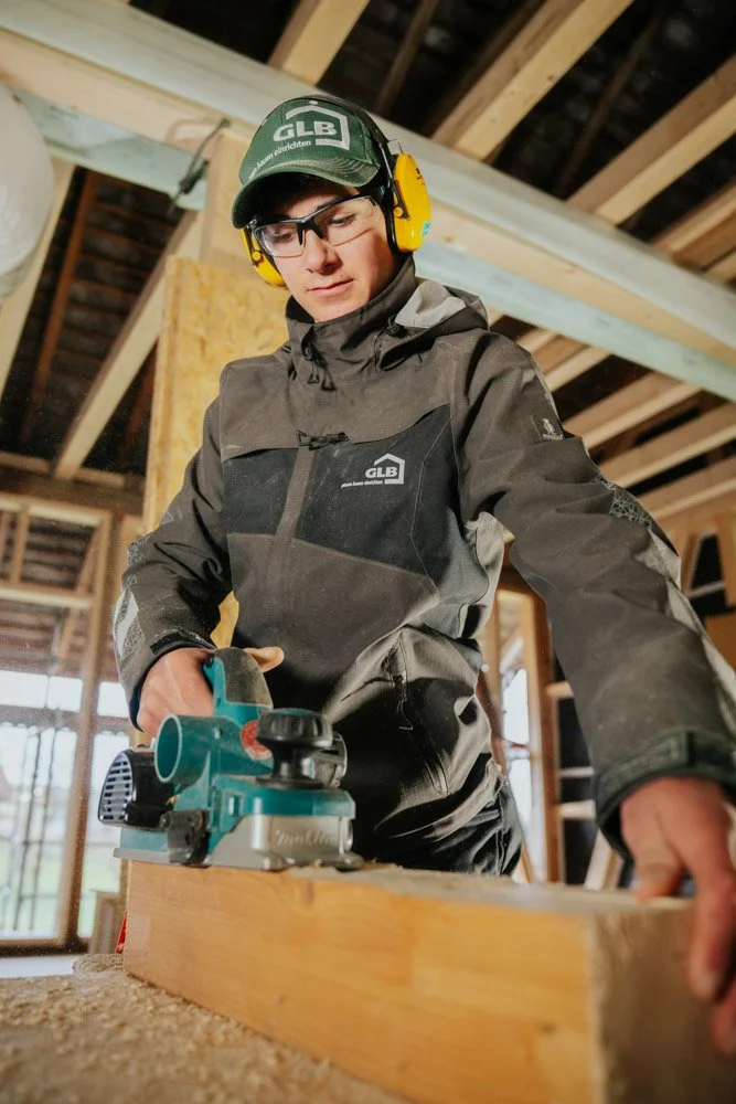Ein junger Mann arbeitet im Holzbau, benutzt eine Handkreissäge an einem Holzbalken, trägt Schutzhelm, Gehörschutz und wetterfeste Kleidung in einem Holzrahmenhaus.