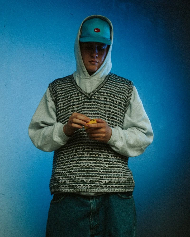 Junge Person trägt eine blaue Kappe, grauen Hoodie und Strickweste, hält Zigarettenpackung in den Händedn, steht vor einer blauen Wand.