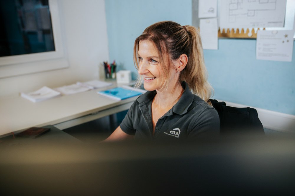 Eine Frau sitzt an einem Schreibtisch mit einem Lächeln, trägt ein schwarzes Polo-Shirt, im Hintergrund sind Arbeitsmaterialien und Poster an der Wand.
