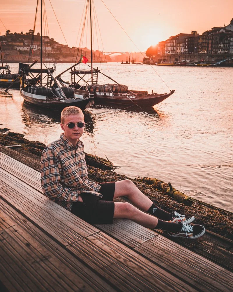 Ein Junge mit Sonnenbrille sitzt an einem Flussufer bei Sonnenuntergang, hinter ihm Boote und eine Stadt.