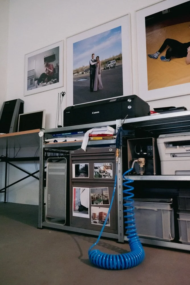 Büro- oder Werkstattbereich mit Drucker, Monitoren, Fotos an der Wand und einem blauen Luftschlauch auf dem Boden.