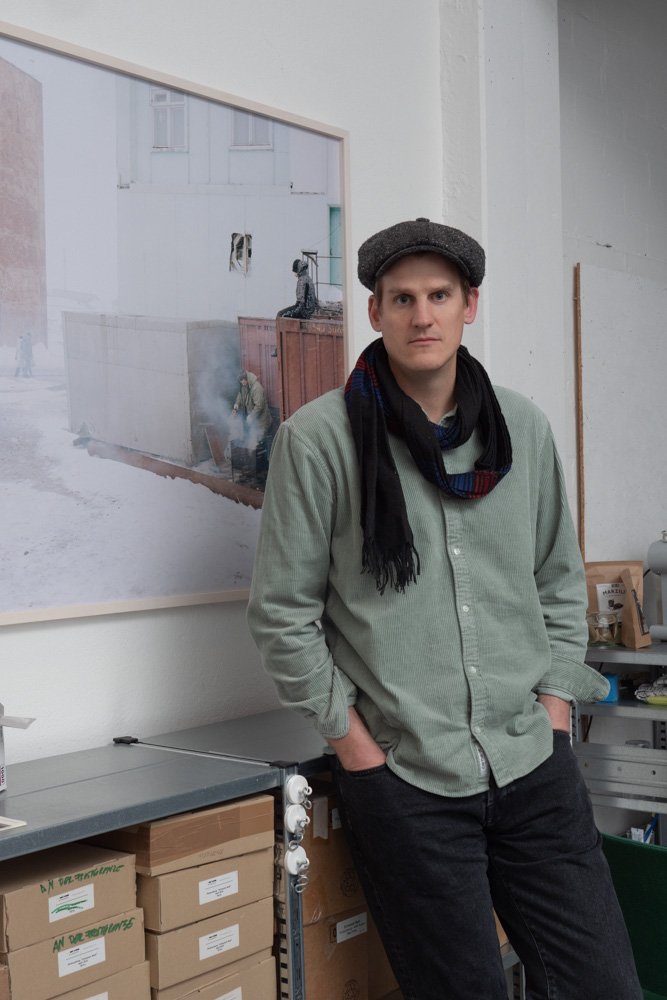 Junger Mann steht in einem Raum vor einem Wandbild, trägt einen grauen Hut, eine grüne Jacke, einen schwarzen Schal und schwarze Hosen, mit den Händen in den Taschen.