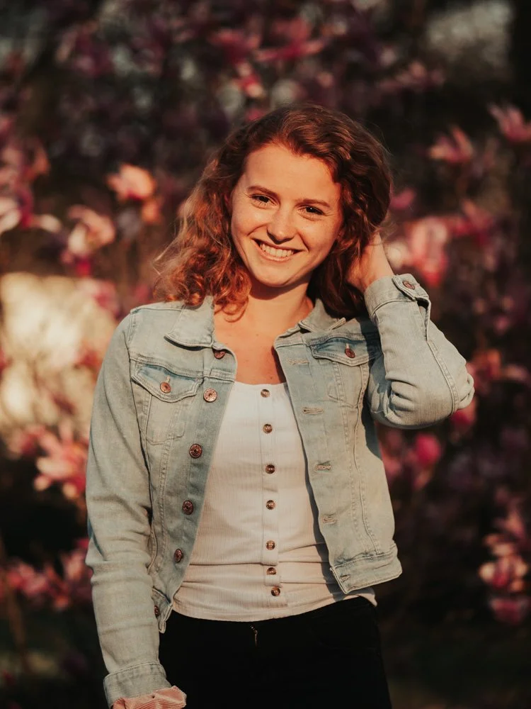 Lächelnde junge Frau mit lockigem braunem Haar, trägt eine Jeansjacke und ein weißes top, steht vor blühenden Kirschblüten im Sonnenuntergang.