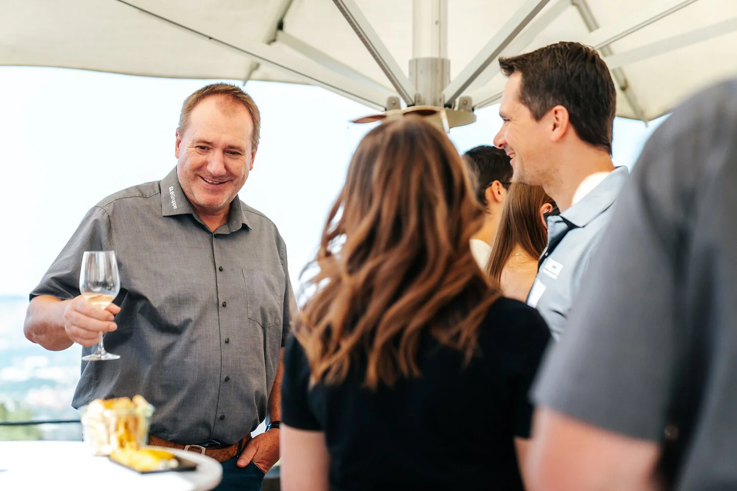 Eine Gruppe von Menschen, die auf einer Party bei sonnigem Wetter unter einem Sonnenschirm stehen, einen Mann mit einem Glas Wein in der Hand.