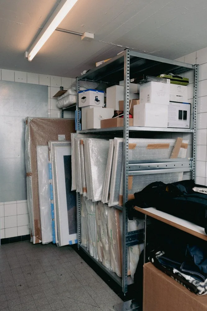 Lagerraum mit Metalregal, gefüllt mit Verpackungsmaterial, gläsernen Objekten, Kartons und einem schwarzen Rucksack auf einem Tisch