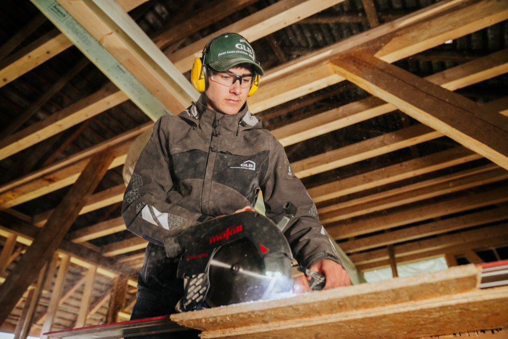 Junge arbeitet mit einer Kreissäge an einer Holzplatte in einem Holzbau, trägt Schutzbrille, Gehörschutz und Arbeitskleidung.