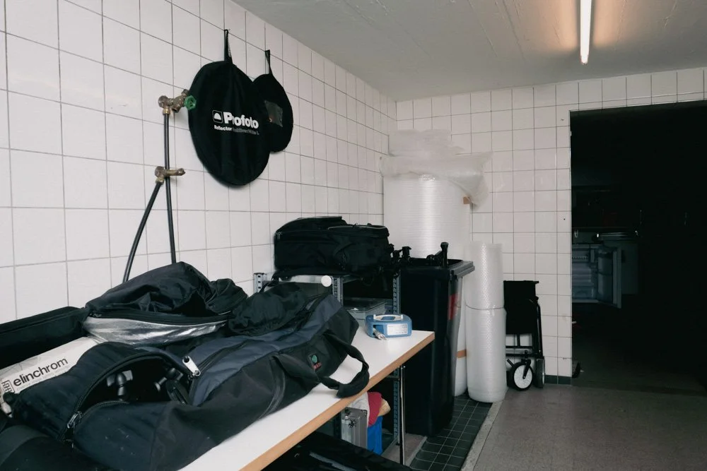 Ein Lagerraum mit weißen Fliesen, auf einem Tisch liegen Kamerataschen, Bags und Equipment sowie Verpackungsmaterial wie Luftpolsterfolie, mehrere Rollen Verpackungsmaterial im Hintergrund, ein Handwagen und eine dunkle Türöffnung.