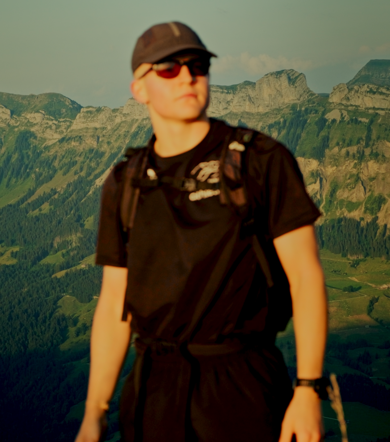 Eine junge Person mit Sonnenbrille, schwarzer Mütze und Rucksack vor einer bergigen Landschaft mit grünen Hügeln und Felsen im Hintergrund.