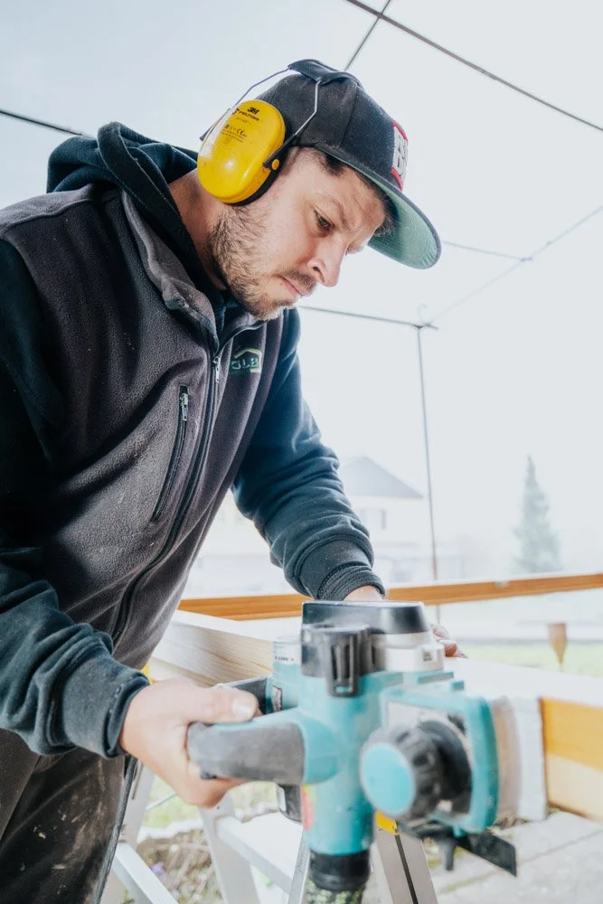 Ein Mann arbeitet mit einem Schleifer an einem Holzbalken, trägt Gehörschutz, eine schwarze Mütze und eine dunkle Jacke.