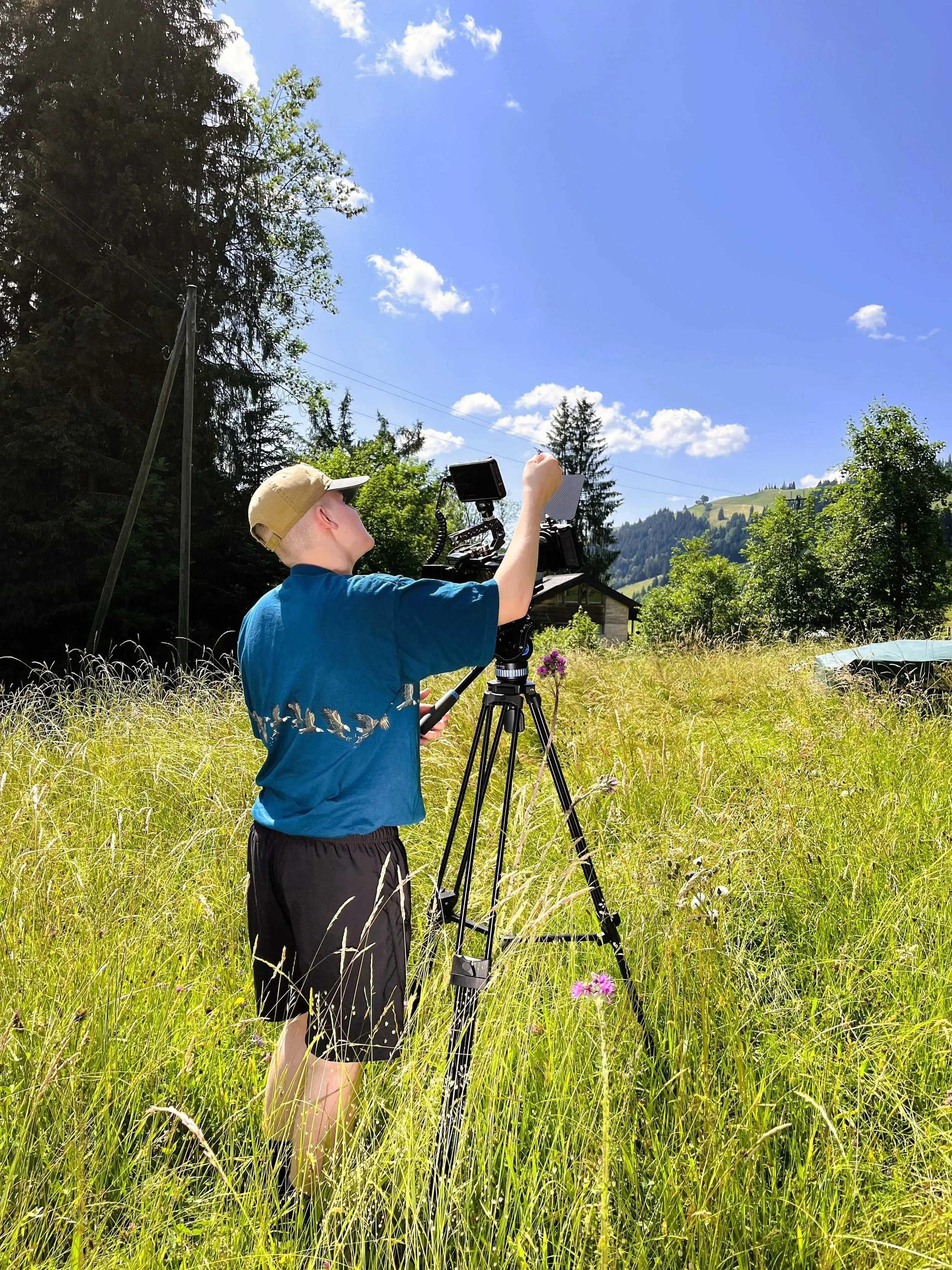 Junge Person, der im Grünen steht, eine Kamera auf einem Stativ bedient, bei sonnigem Wetter in einer ländlichen Umgebung mit Bäumen, Hügeln und einem blauen Himmel.