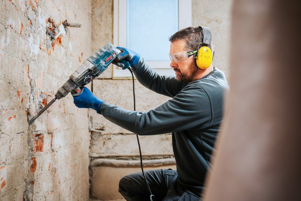 Mann mit einem Bohrhammer, trägt Schutzbrille, Gehörschutz und Handschuhe.
