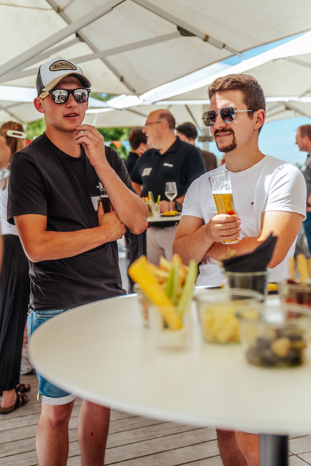 Zwei junge Männer unter einem Sonnenschirm im Gespräch, einer hält ein Glas Bier, im Hintergrund andere Gäste bei einer Feier