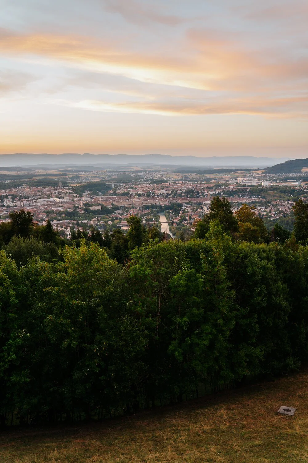 Blick über eine Stadt mit Fluss im Stadtzentrum, umgeben von Hügeln und einer bewaldeten Gegend bei Sonnenuntergang.