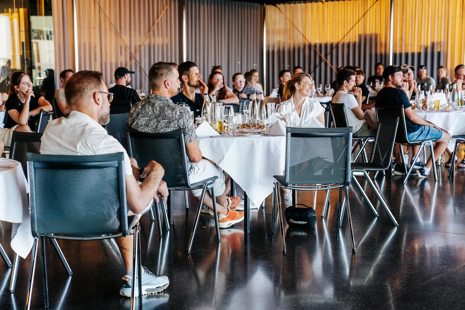 Menschen sitzen bei einer Veranstaltung an runden Tischen im Restaurant, einige schauen nach vorne, andere setzen sich entspannt in einem hell erleuchteten Raum mit großen Fenstern und Vorhängen.