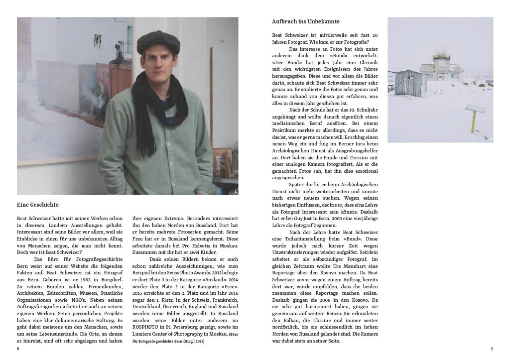 Eine Person steht in einem Raum vor einer Wand mit Kunstwerken, trägt eine grüne Jacke, eine dunkle Mütze und einen Schal.