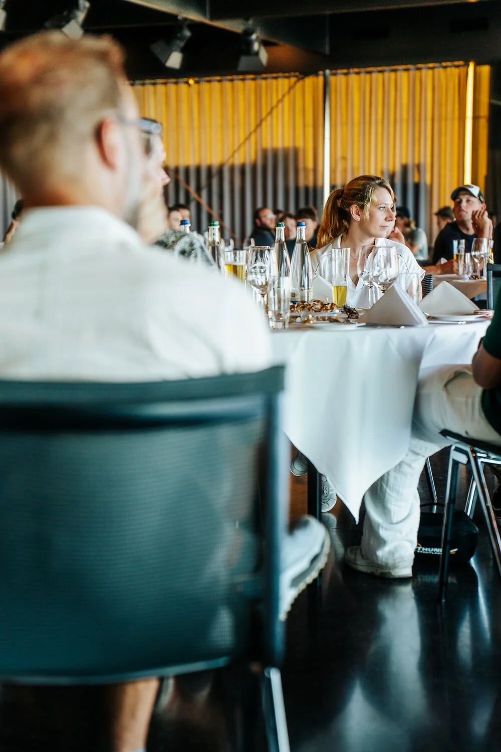 Menschen sitzen an einem gedeckten Tisch in einem Restaurant oder Veranstaltungsraum, trinken und hören aufmerksam zu.