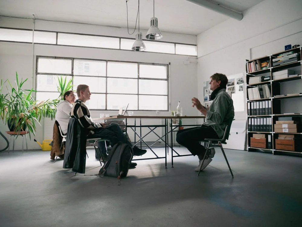 Drei junge Menschen sitzen an einem Tisch, während eine Frau einem Mann gegenüber spricht, wahrscheinlich im Rahmen eines Bewerbungsgesprächs oder Meetings, in einem modernen, gut beleuchteten Büro mit Pflanzen, Büchern und Regalen.