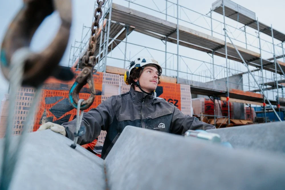 Bauarbeiter mit Helm auf einer Baustelle, im Hintergrund Gerüst und Baumaterialien.
