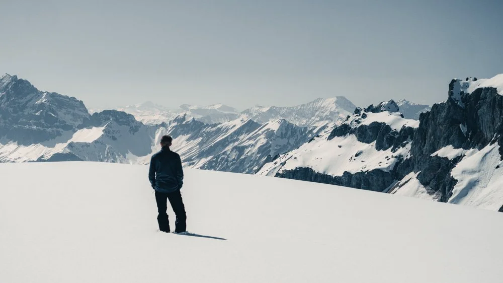 Ein Mensch steht in einer schneebedeckten Berglandschaft mit hohen, schneebedeckten Gipfeln und dunklen Felsen im Hintergrund.