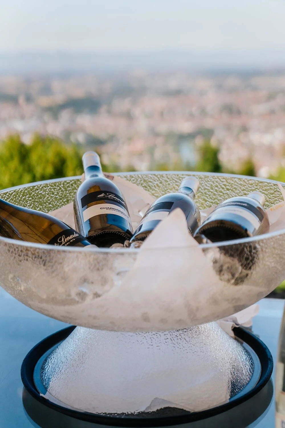 Mehrere Champagnerflaschen in einem Eiskübel mit Eiswürfeln, auf einem Tisch vor einer Stadt- und Landschaftsansicht an einem sonnigen Tag.