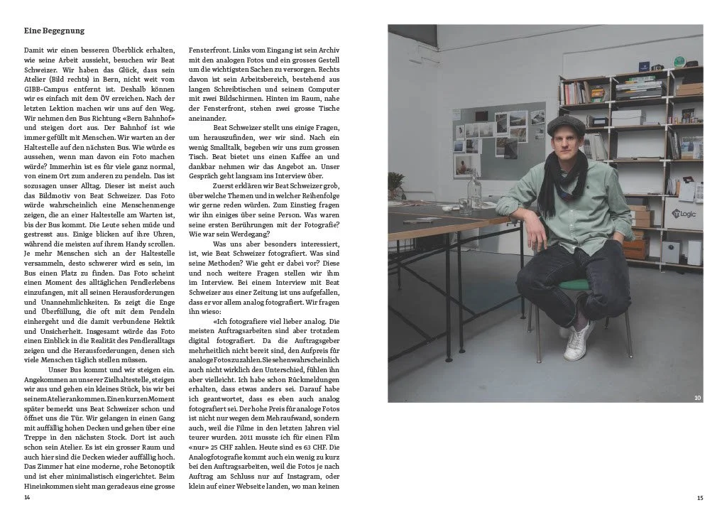 Ein junger Mann sitzt in einem Bücherregalraum. Er trägt eine helle Jacke, eine schwarze Kappe und weiße Schuhe. Auf dem Tisch vor ihm liegen zwei Computer und Notizen. Der Raum wirkt modern und gut ausgestattet.