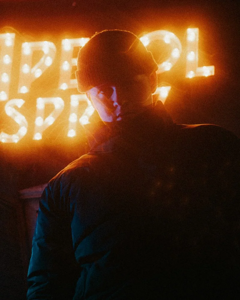 Eine Person mit Mütze steht vor leuchtenden Buchstaben, die das Wort 'APEROL SPRITZ' bilden, in einem dunklen Ambiente.