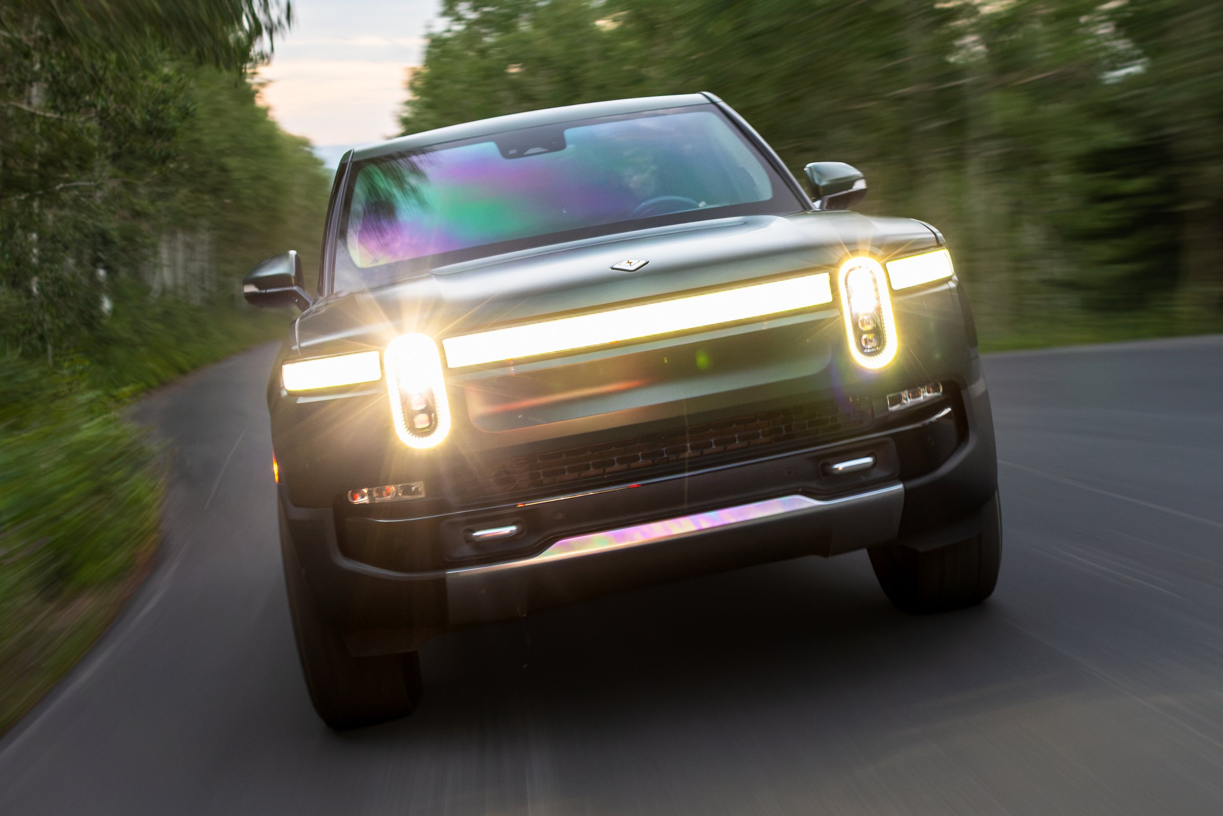 A green Rivian R1T truck with illuminated headlights and taillights, driving on a winding road through a green forest during sunset.