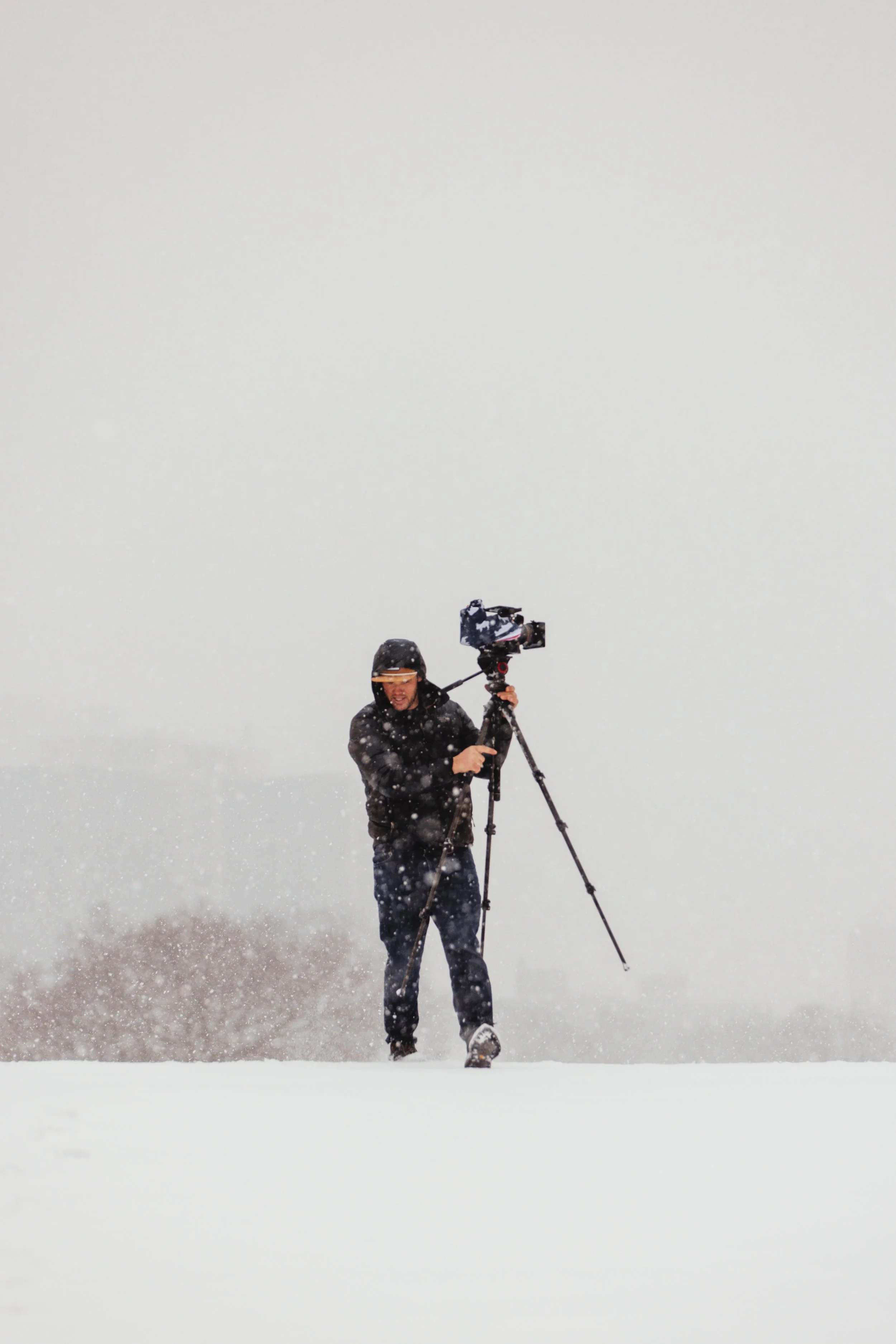 A person wearing a black jacket, pants, and gloves, standing in a snowy landscape while operating a RED camera on a tripod during a snowstorm.