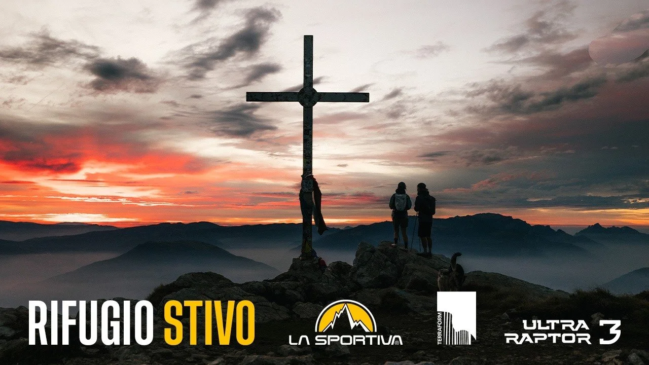 Two hikers standing on a mountain at sunset near a cross-shaped monument, with misty mountains in the background. Text logos show Rifugio Stivo, La Sportiva, Terraform Creative, and Ultra Raptor III.