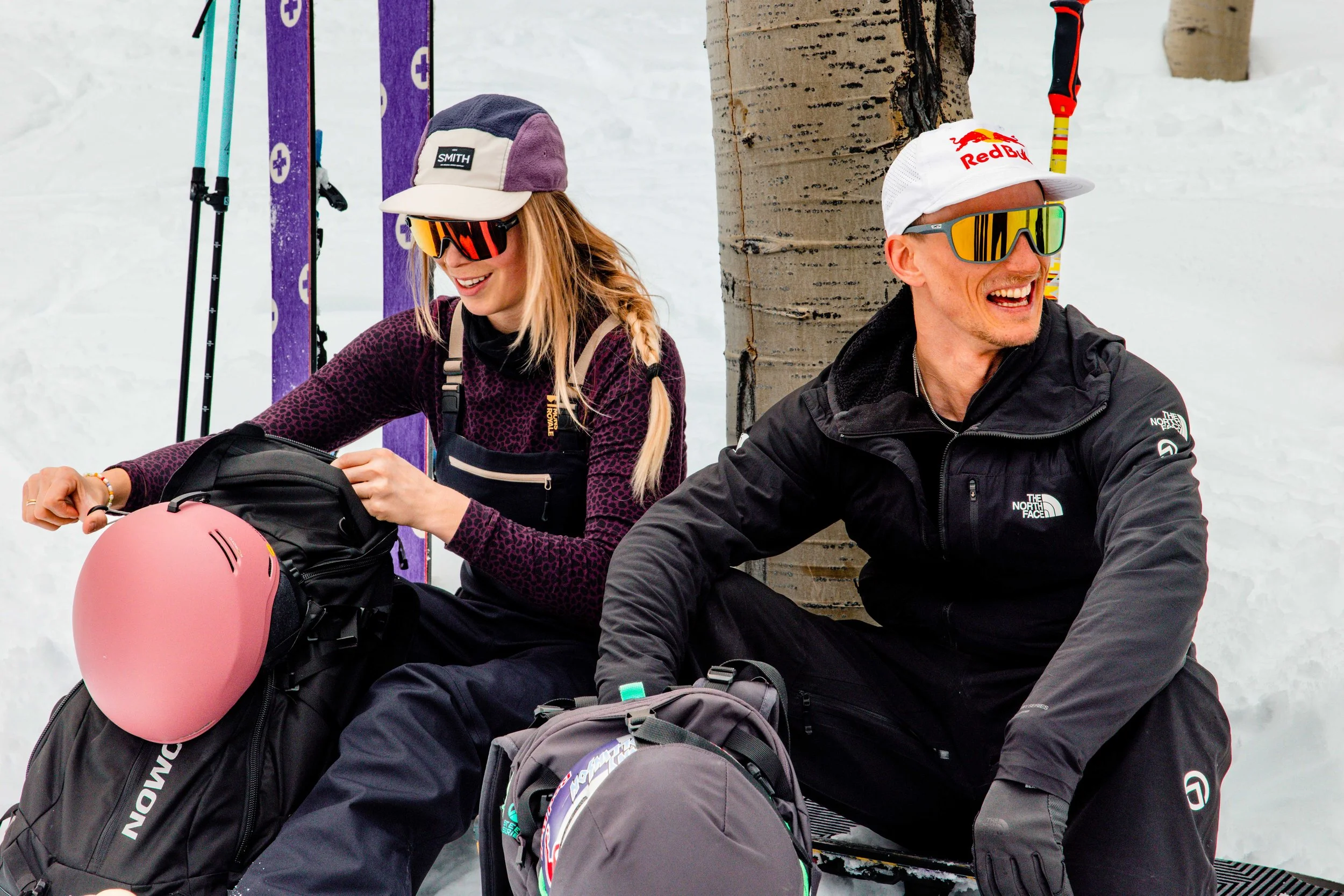 Two people sitting on snow, wearing ski gear and sunglasses, smiling, with skiing equipment nearby.