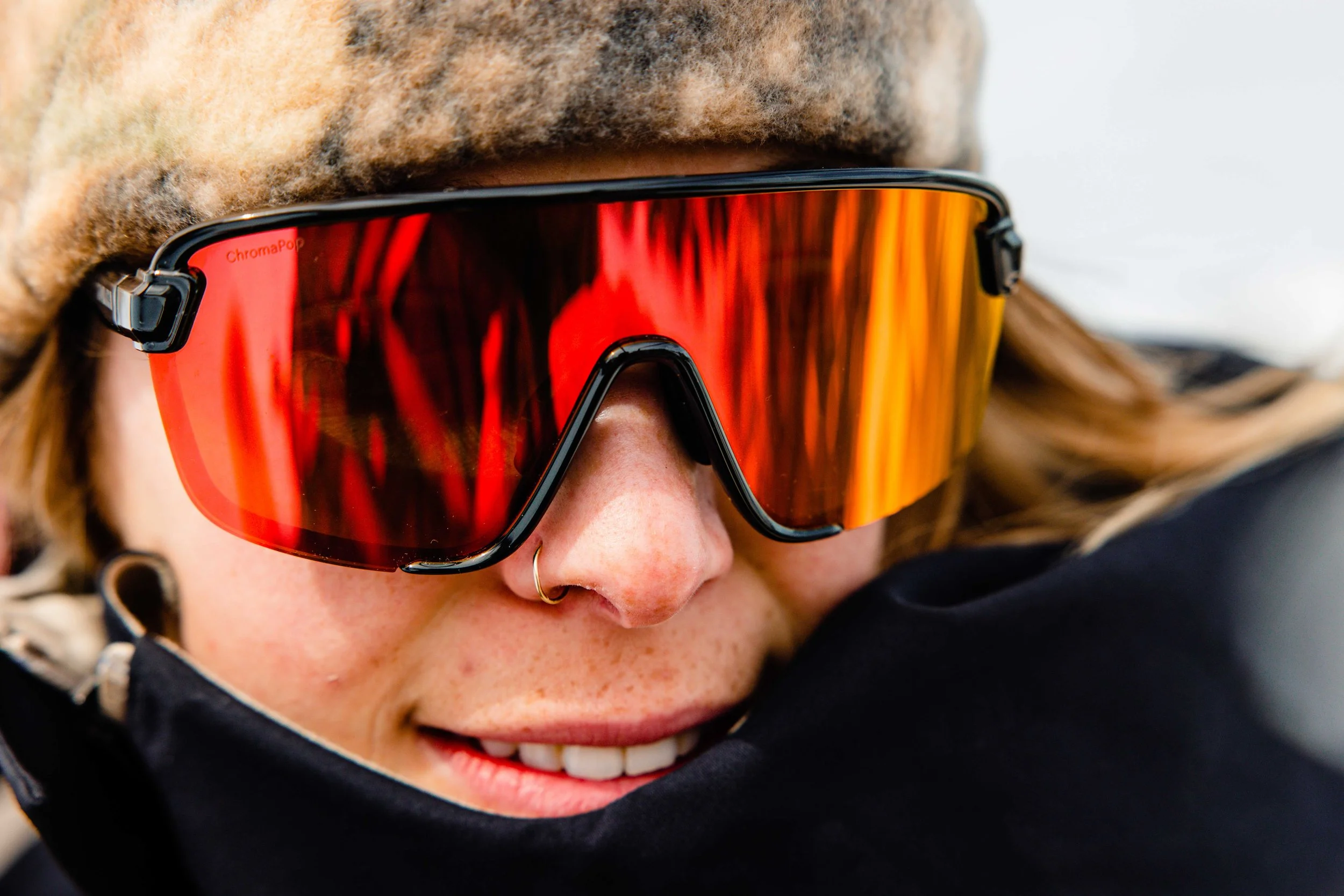 Close-up of a person wearing large, orange-tinted Smith Optics sports sunglasses, a furry hat, and a nose ring, smiling with visible teeth.