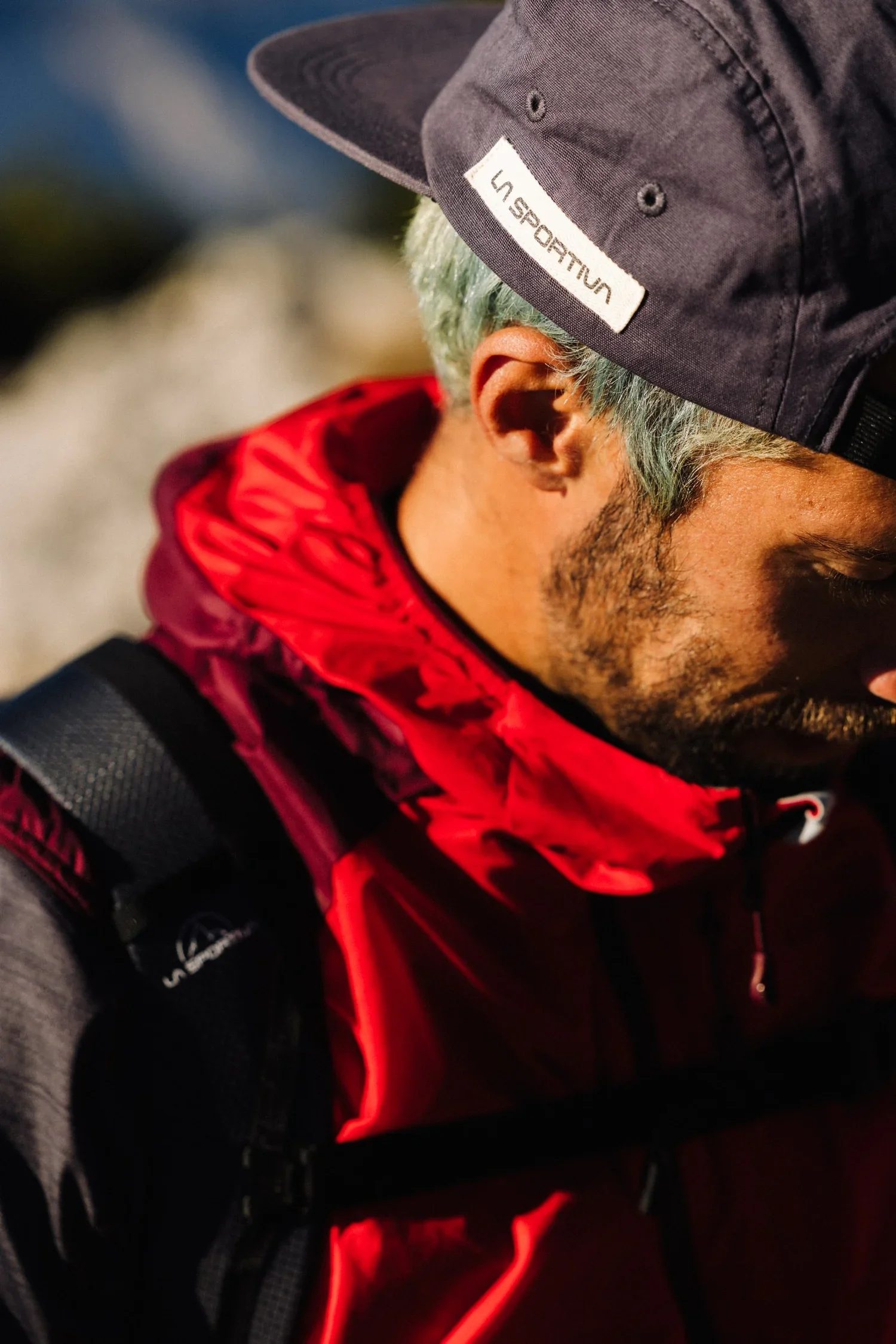 Close-up of a person wearing a black cap with a "La Sportiva" logo, a red jacket, and a black backpack with red accents, outdoors at night.