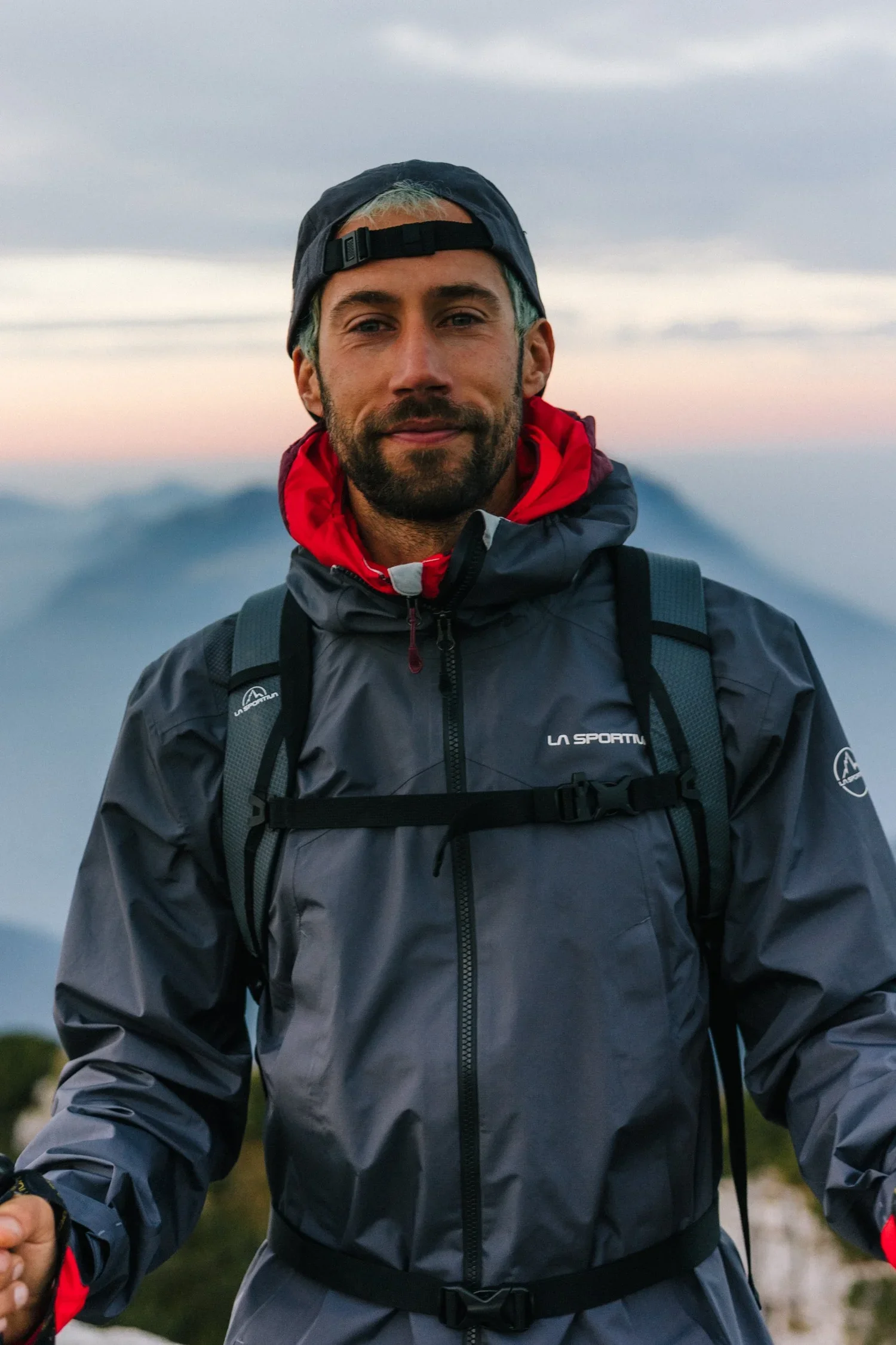 A man with a beard wearing a gray La Sportiva jacket, a backwards baseball cap, and a backpack, standing outdoors during sunset with mountains and a cloudy sky in the background.