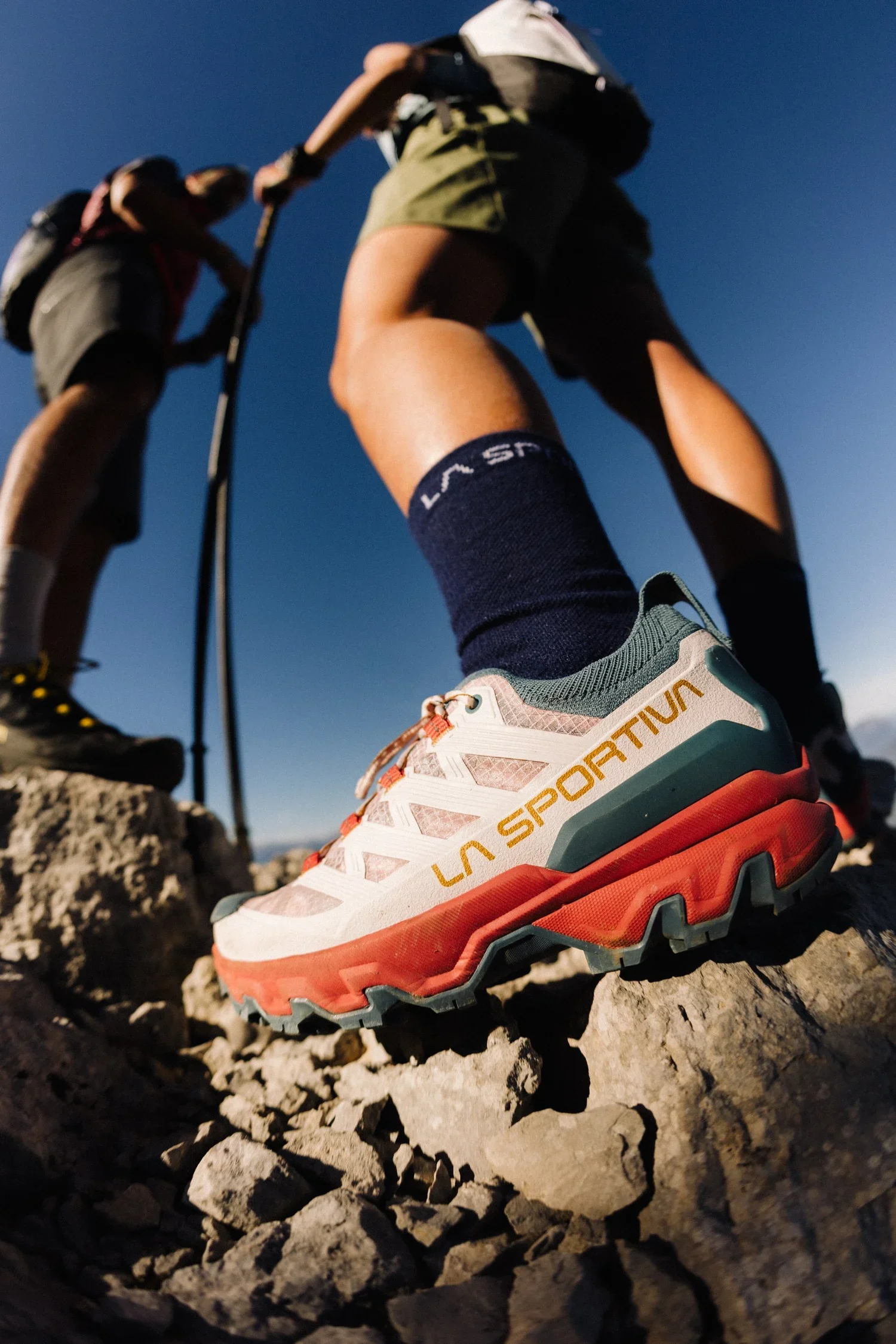 Close-up of a hiker's sneaker on rocky terrain, with two hikers in the background on a mountain trail under a clear blue sky.