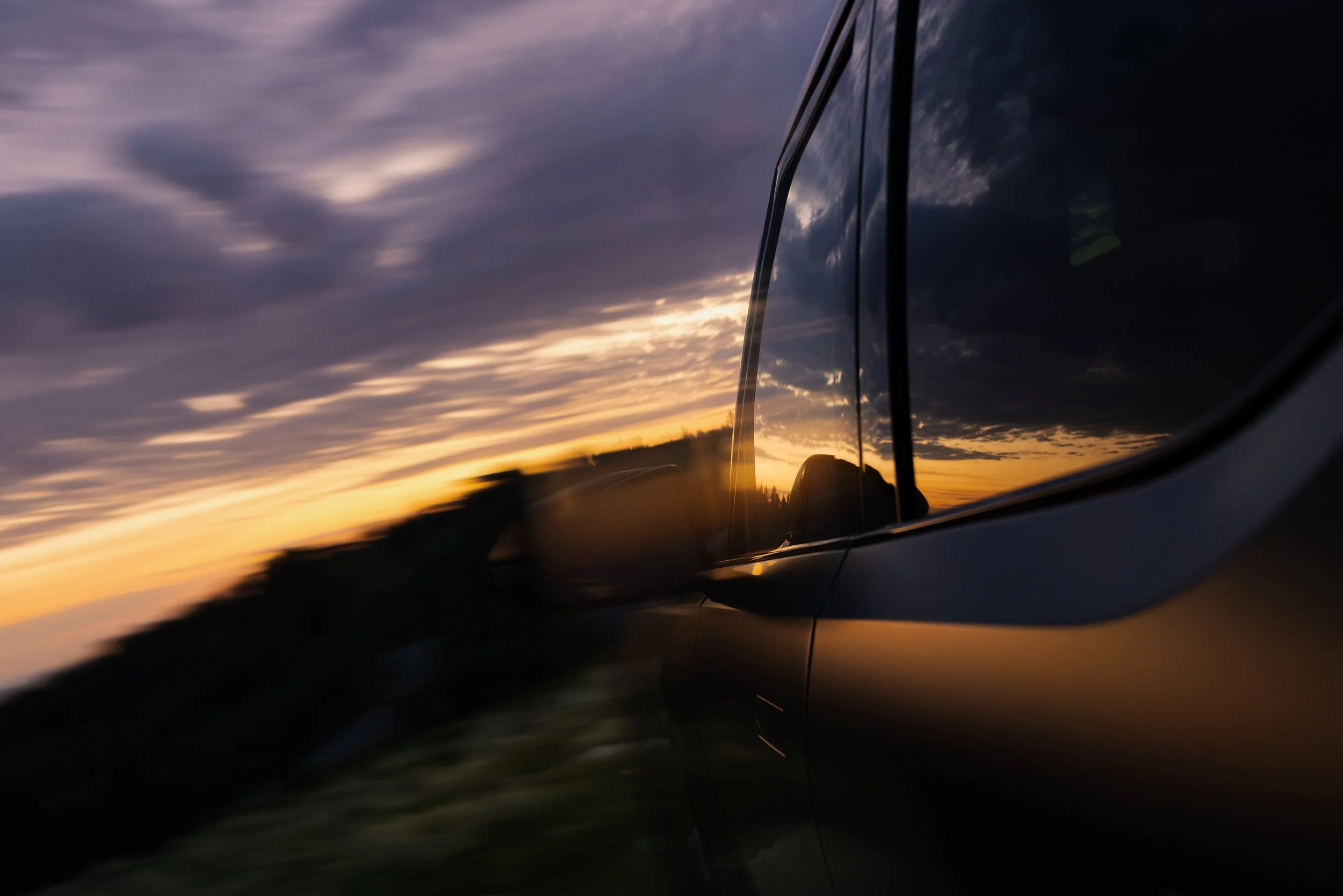 The photo shows a sunset with orange and purple sky reflections on a car's side mirror, capturing both the sky and part of the vehicle in motion.