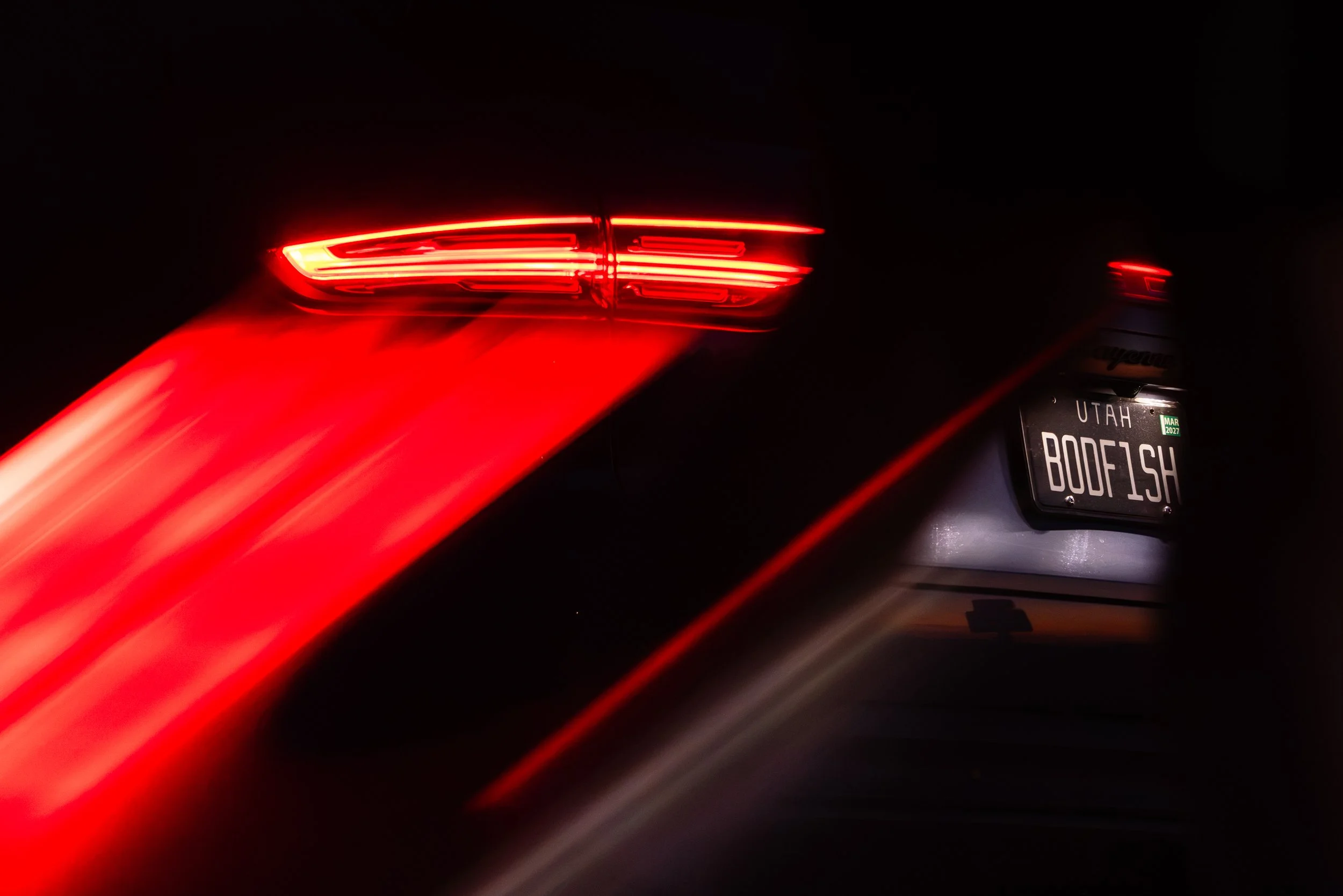 Close-up of a red car's tail light at night with motion blur. The vehicle has a Utah license plate reading 'BODFISH' and a sticker indicating March 2027 expiration date.