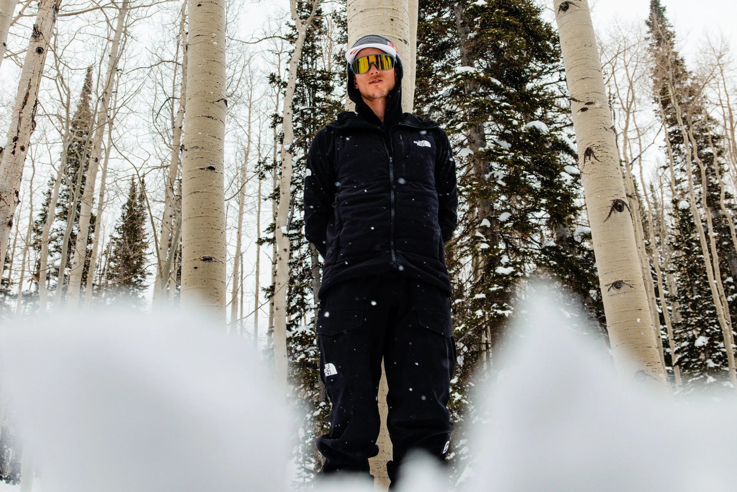 John Collinson dressed in ski gear, including a black jacket, black pants, ski goggles, and a white hat, stands among tall, white-barked aspen trees in a snowy forest with snow-covered evergreen trees in the background.