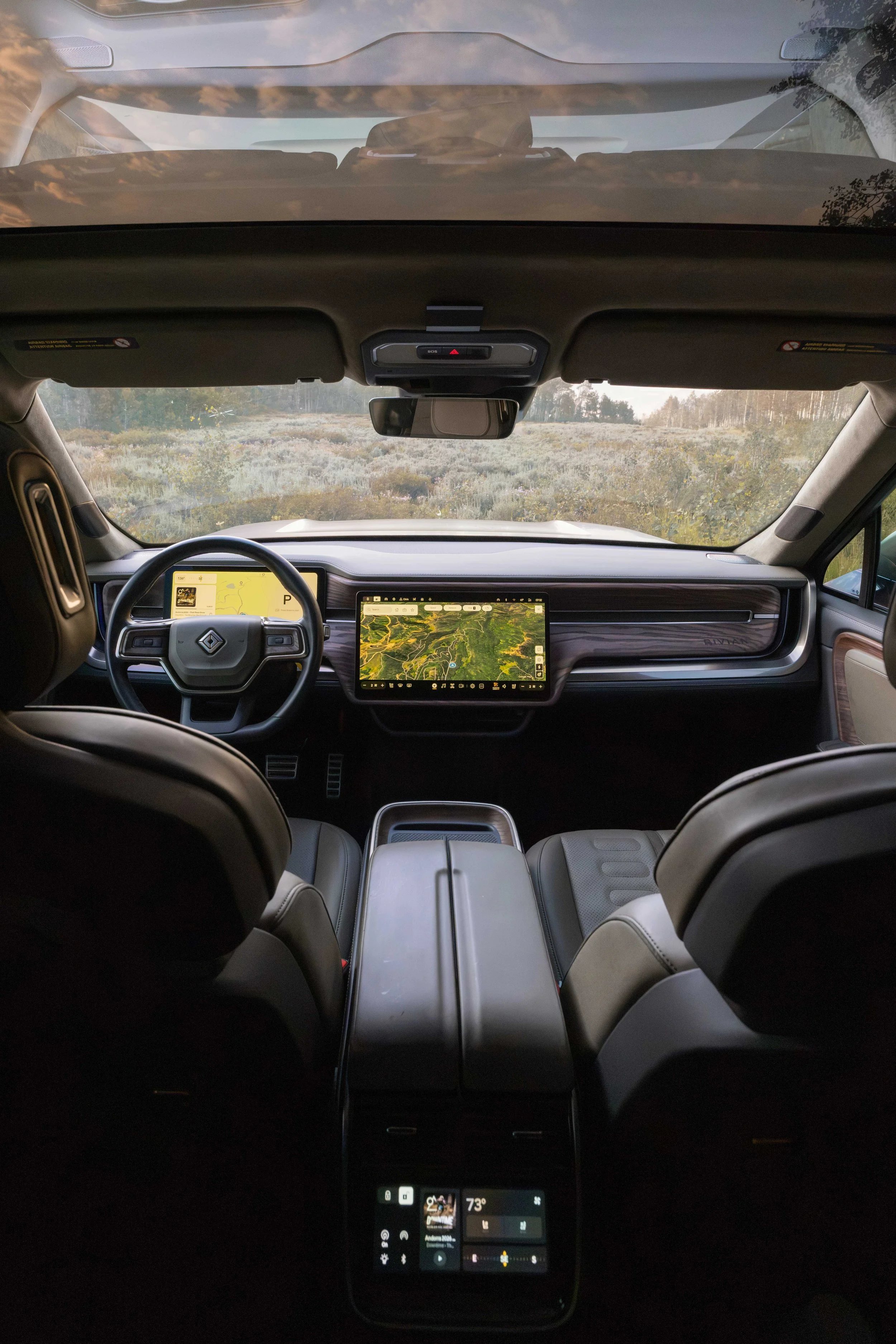 Interior view of a Rivian R1T vehicle showing a dashboard with two large screens, steering wheel, and front seats, looking out onto a landscape of trees and open land through the windshield at dusk or dawn.