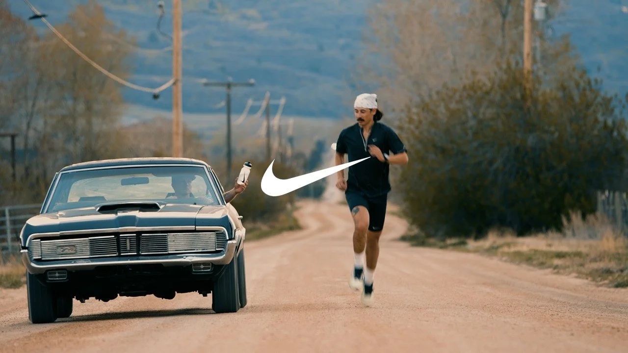 A man in running clothes jogging on a dirt road while a car pulls alongside him, passing a hand holding a water bottle. The Nike logo is on screen.