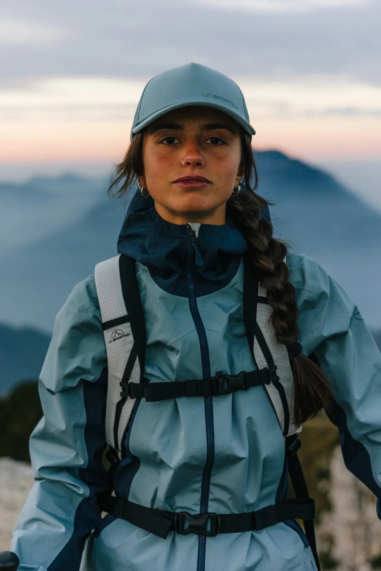 A woman with long braided hair wearing a gray cap, a blue jacket, and a backpack outdoors during sunset or sunrise with mountains in the background.