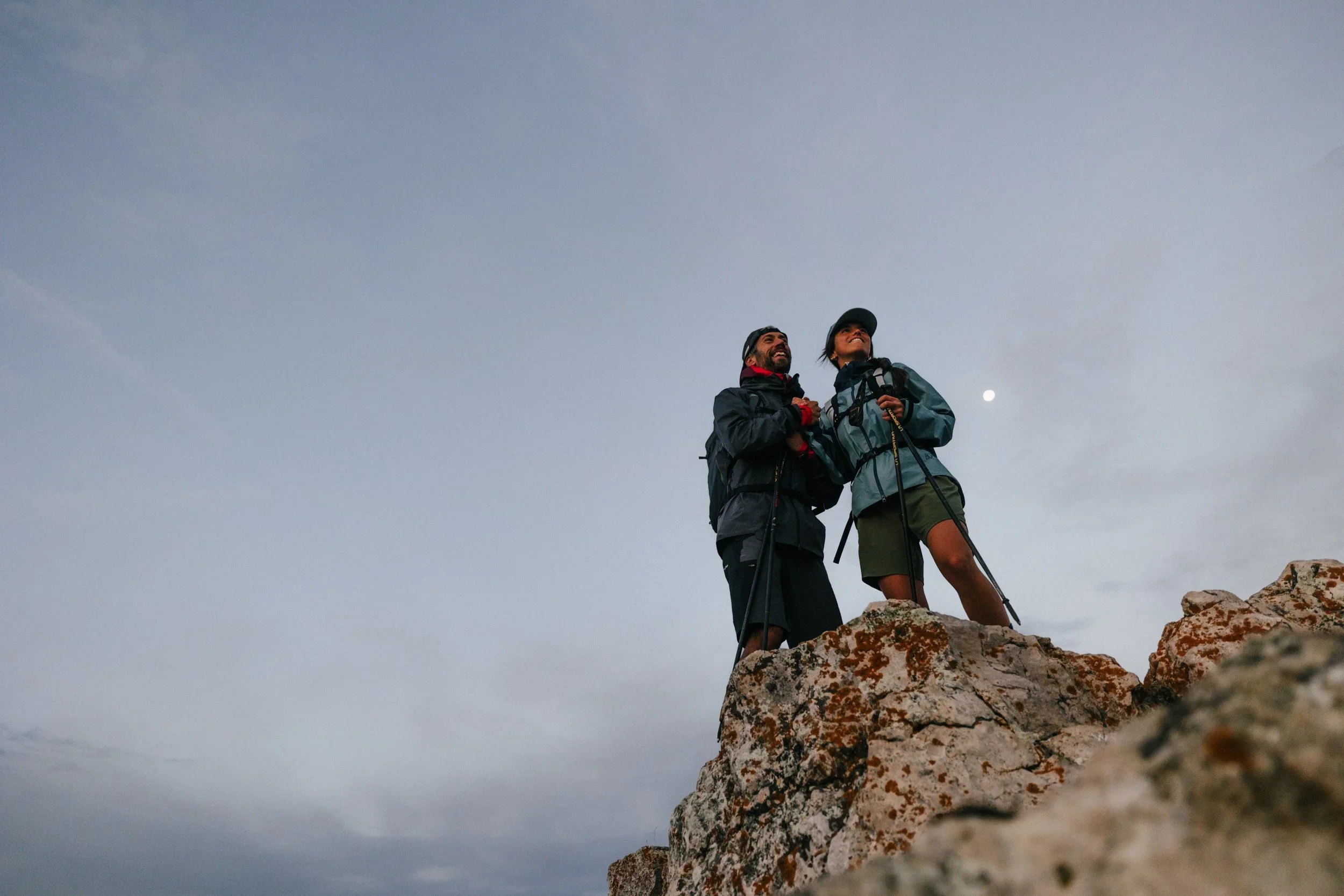 Two hikers standing on a rocky cliff, looking up at the sky, with the moon visible in the background during dusk or dawn.