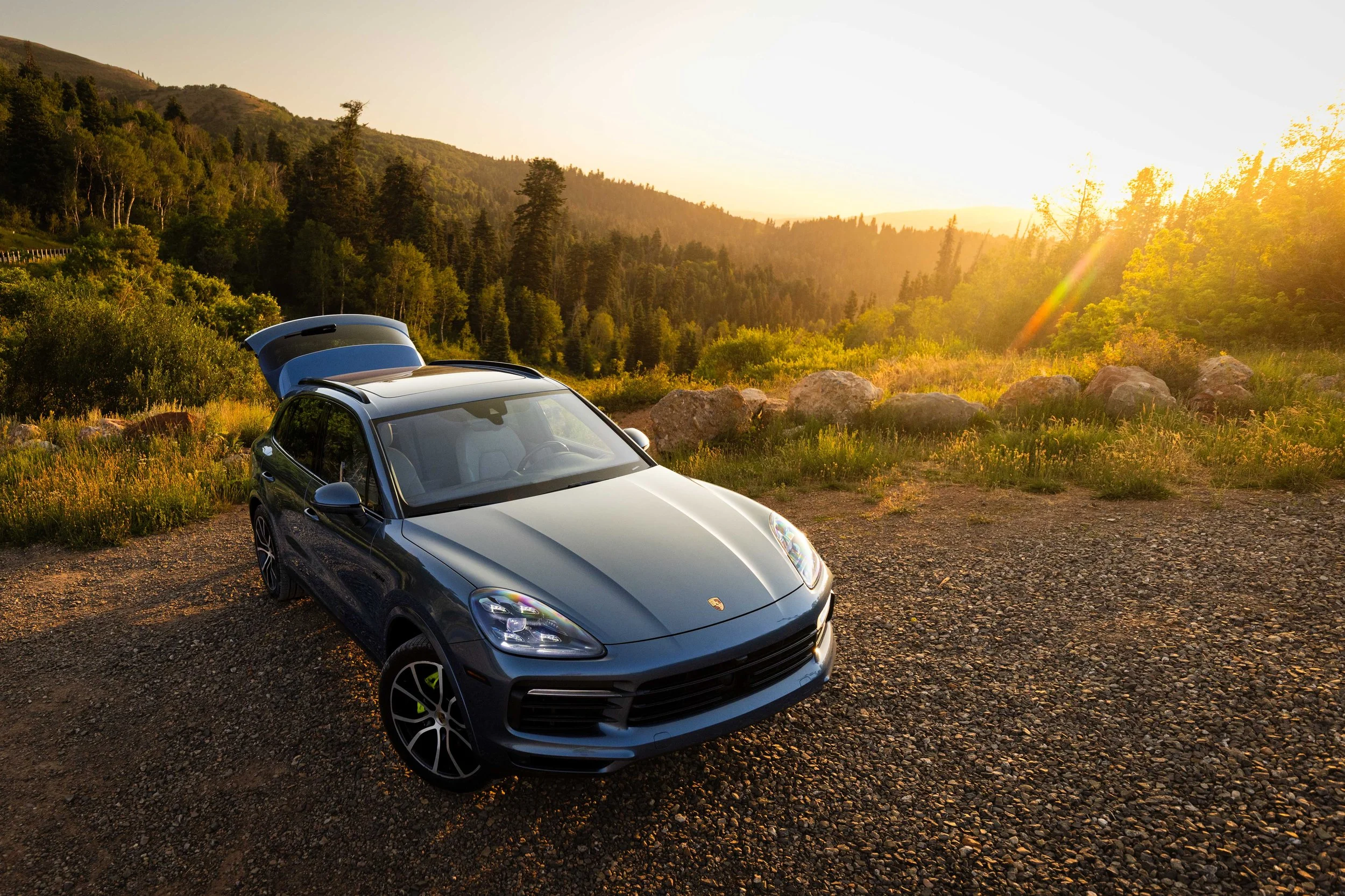 A sleek gray Porsche SUV parked on a dirt clearing overlooking a lush green forested mountainside during sunset, with sunlight casting a warm golden glow over the scene.